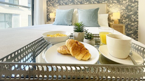 Ein ordentlich gedecktes Frühstücksbrettchen mit Croissants, einer Schüssel Müsli, einer Tasse und einem Glas Orangensaft auf einem Glastisch in einem Hotelzimmer mit Bett, Kissen und floralem Wandpapier.