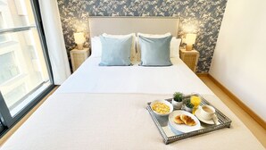 Ein ordentlich bezogenes Bett mit zwei Kissen, ein Tablett mit Frühstücksangeboten und ein Fenster mit Blick auf Gebäude.