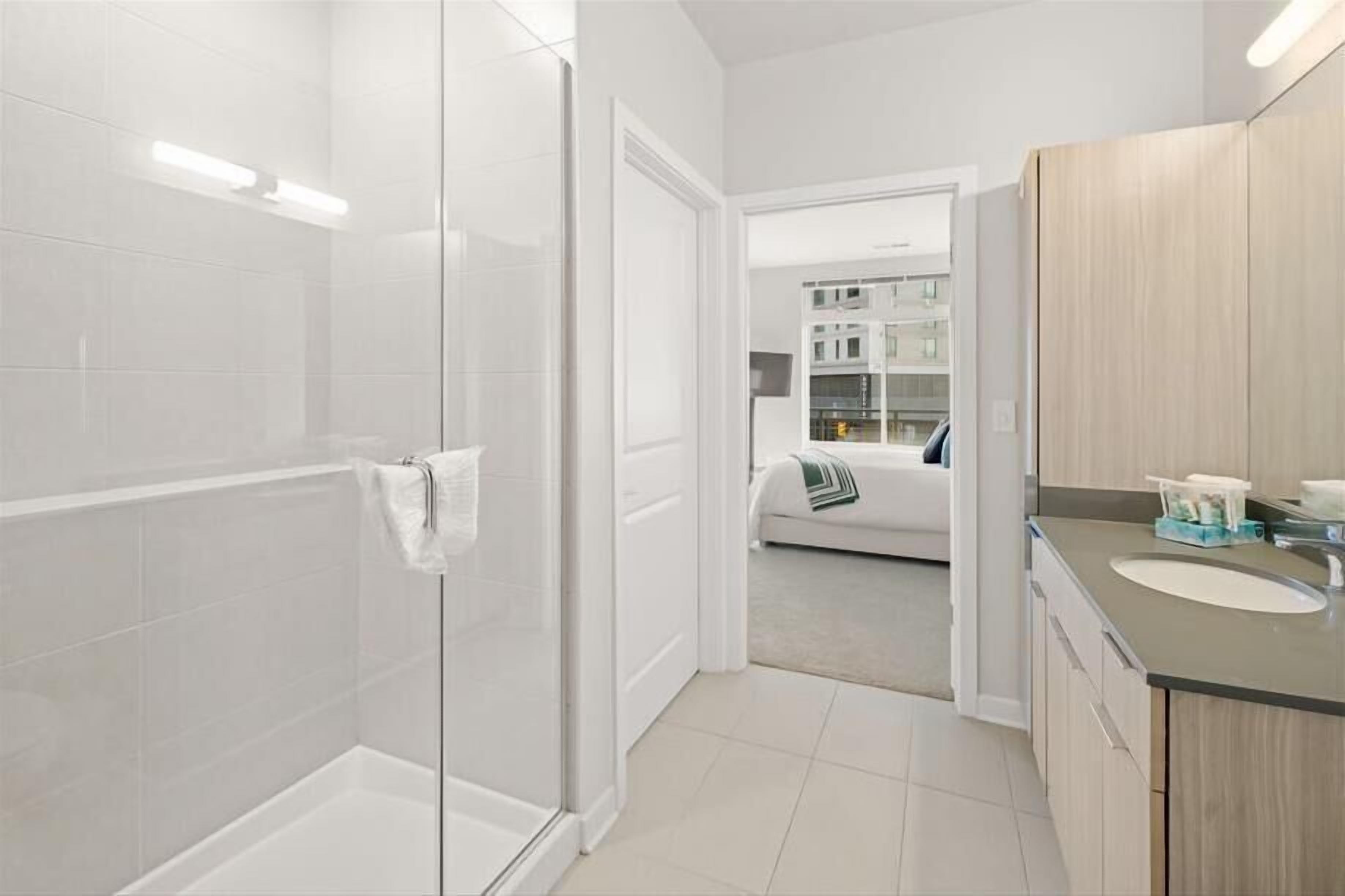 A modern bathroom with a glass shower enclosure, a sink with a wooden vanity, and a visible bedroom with a bed and a nightstand.