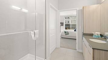 A modern bathroom with a glass shower enclosure, a sink with a wooden vanity, and a visible bedroom with a bed and a nightstand.
