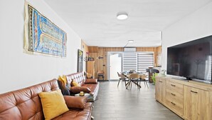 Een moderne woonkamer met een leren bank, houten kasten, een flatscreen televisie en een eethoek met stoelen en een tafel.
