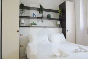 A hotel room with a bed, white linens, a bedside table with a lamp, and shelves with plants and a letter 'b'. - Homiday - Felicioni Alba, 2-room apartment on the 1st floor (Alba Adriatica)