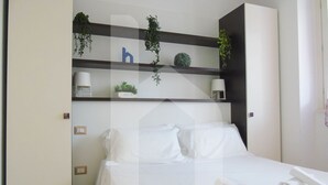 A hotel room with a bed, white linens, a bedside table with a lamp, and shelves with plants and a letter 'b'. - Homiday - Felicioni Alba, Two-room apartment on the 1st Floor (Alba Adriatica)