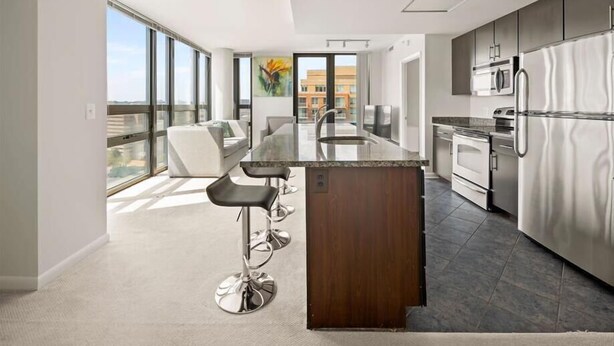 A modern kitchen with stainless steel appliances, a central island with bar stools, and large windows offering a city view. - Crystal City 2 BR | 10 min drive to Capitol (Arlington)