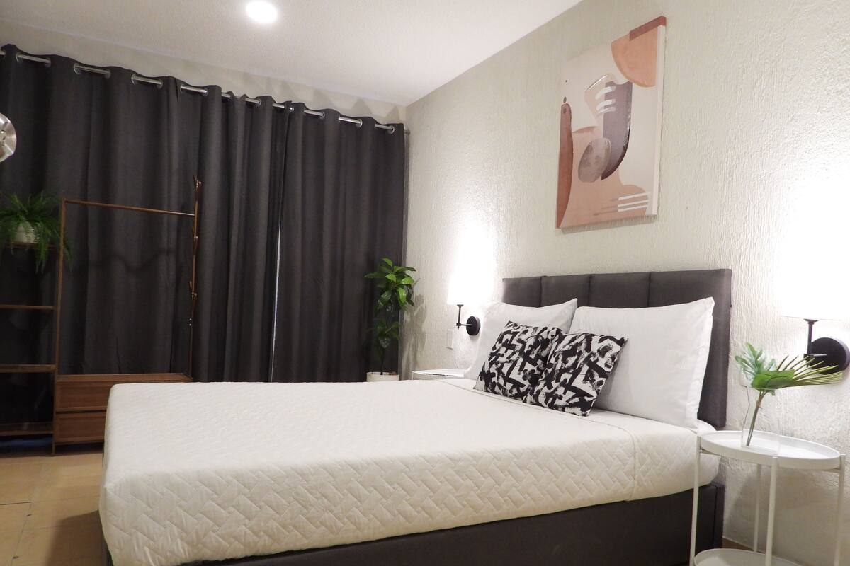 Una cama bien tendida con un edredón blanco acolchado, un cabecero oscuro y dos almohadas estampadas. Hay una mesita de noche blanca con una lámpara, y una planta en maceta en el suelo al lado de la cama.