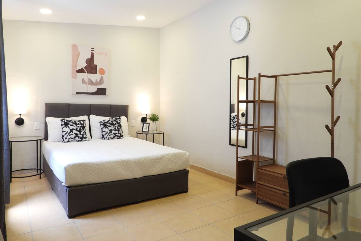 Un dormitorio moderno con cama, mesitas de noche, espejo, reloj y perchero.