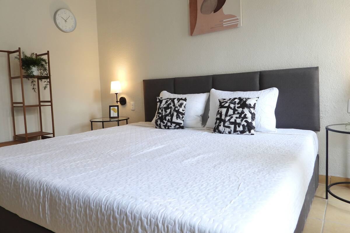 Una cama bien hecha con un edredón blanco con textura, un cabecero oscuro y dos almohadas estampadas. Se observa un reloj de pared y un cuadro enmarcado en la pared.