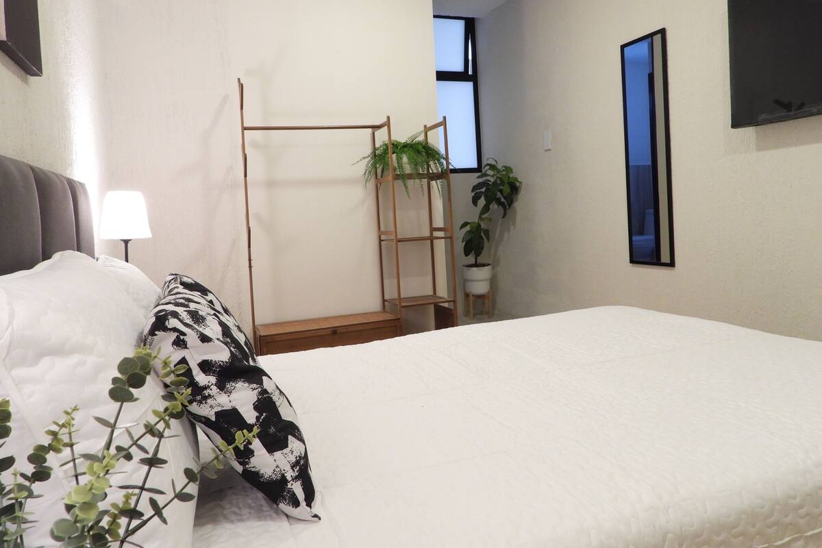 Un dormitorio con una cama, una mesita de noche con lámpara, una escalera de madera y dos plantas en macetas.