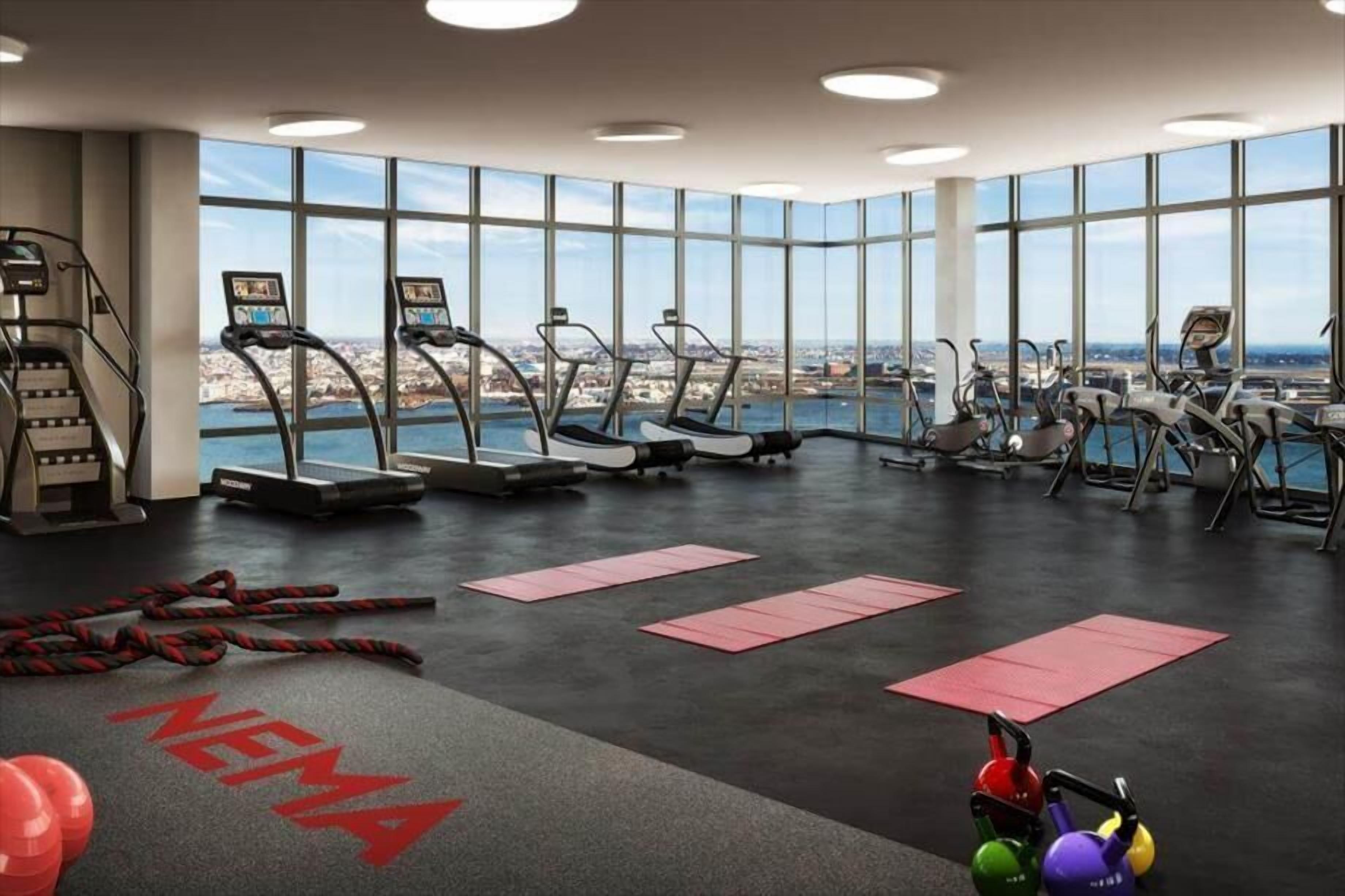 Un gimnasio con cintas de correr, una escalera y vistas a un paisaje urbano a través de amplios ventanales.