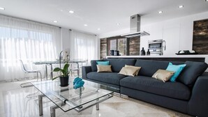 Ein modernes Wohnzimmer mit einem Glastisch, einem dunkelgrauen Sofa mit vielen Kissen und einer Küchenzeile inklusive Herd und Backofen.