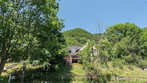 Une maison avec un escalier menant à une terrasse, entourée d’une végétation luxuriante et d’arbres.