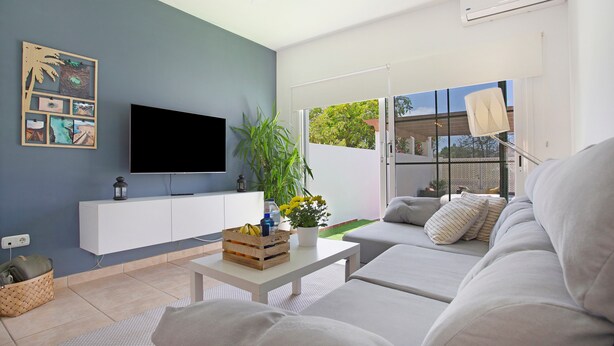 A modern living room with a flat-screen TV, a white sofa, a coffee table with a fruit basket, and a potted plant.