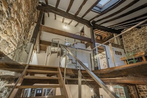 Et rustikt interiør med synlige stenvægge, træbjælker og en trappe med glasgelændere.
