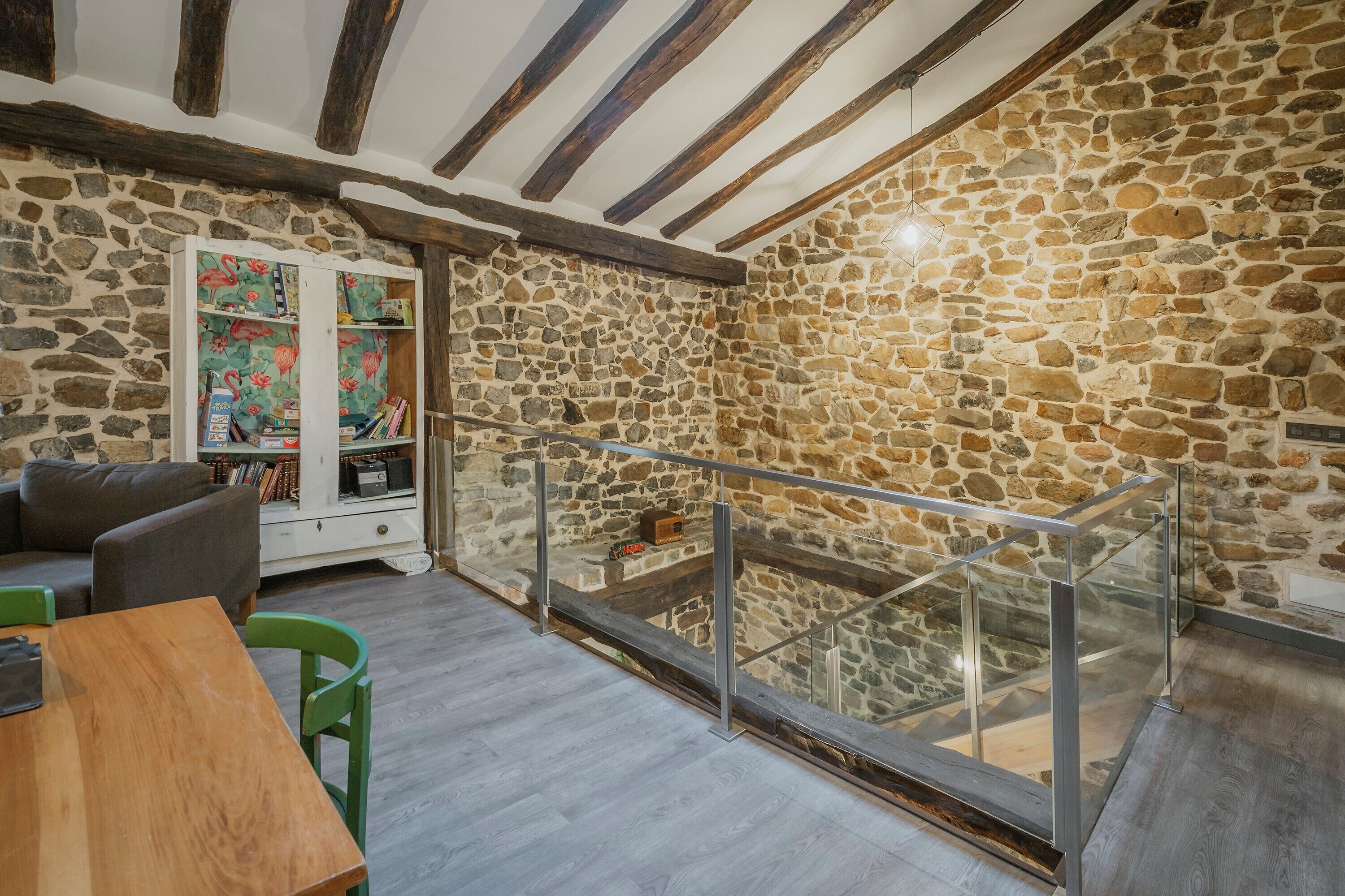 A room with a stone wall, wooden beams, a glass railing, a sofa, a table, and a bookshelf.