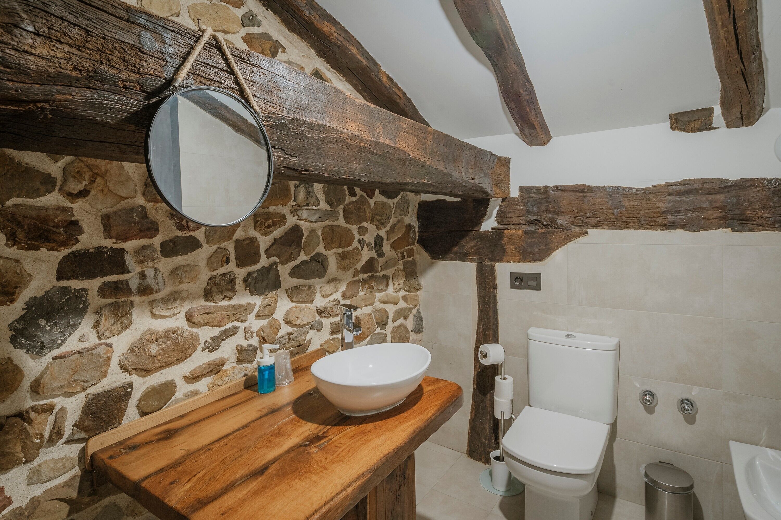 Un baño con pared de piedra, techo con vigas de madera, encimera de madera, lavamanos blanco y retrete blanco.