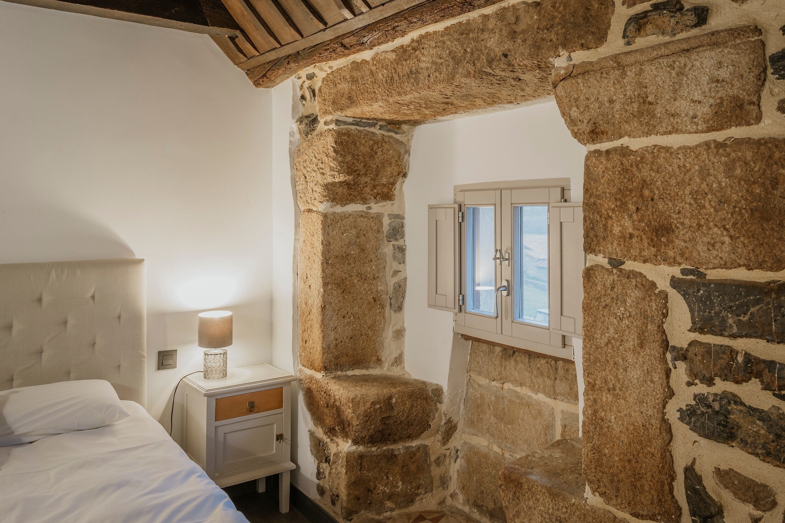 Un dormitorio con pared de piedra, una cama con ropa de cama blanca, una mesita de noche con una lámpara y una ventana con contraventanas de madera.