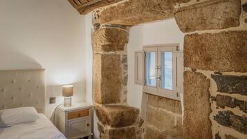 Un dormitorio con pared de piedra, una cama con ropa de cama blanca, una mesita de noche con una lámpara y una ventana con contraventanas de madera.