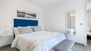 Un dormitorio con cama, una mesita de noche con lámpara, un banco, un espejo grande y un cuadro colgado en la pared.