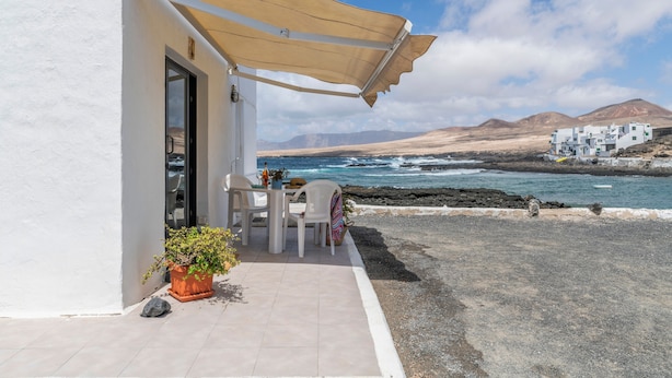 Un edificio blanco con un patio, una mesa, sillas, una planta en maceta y vista al mar.