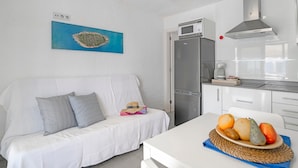 Un sofá blanco con almohadones, una zona de cocina con refrigerador, microondas y estufa, y un frutero sobre una mesa.
