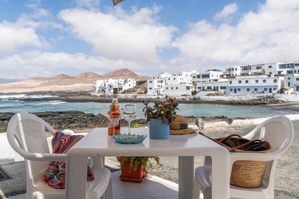 A table with a bottle, glasses, and a plant, overlooking a coastal village with white buildings. - Holiday Home 'La Casita Del Mar' Seafront, Private Terrace & Wi-Fi (Caleta de Caballo)