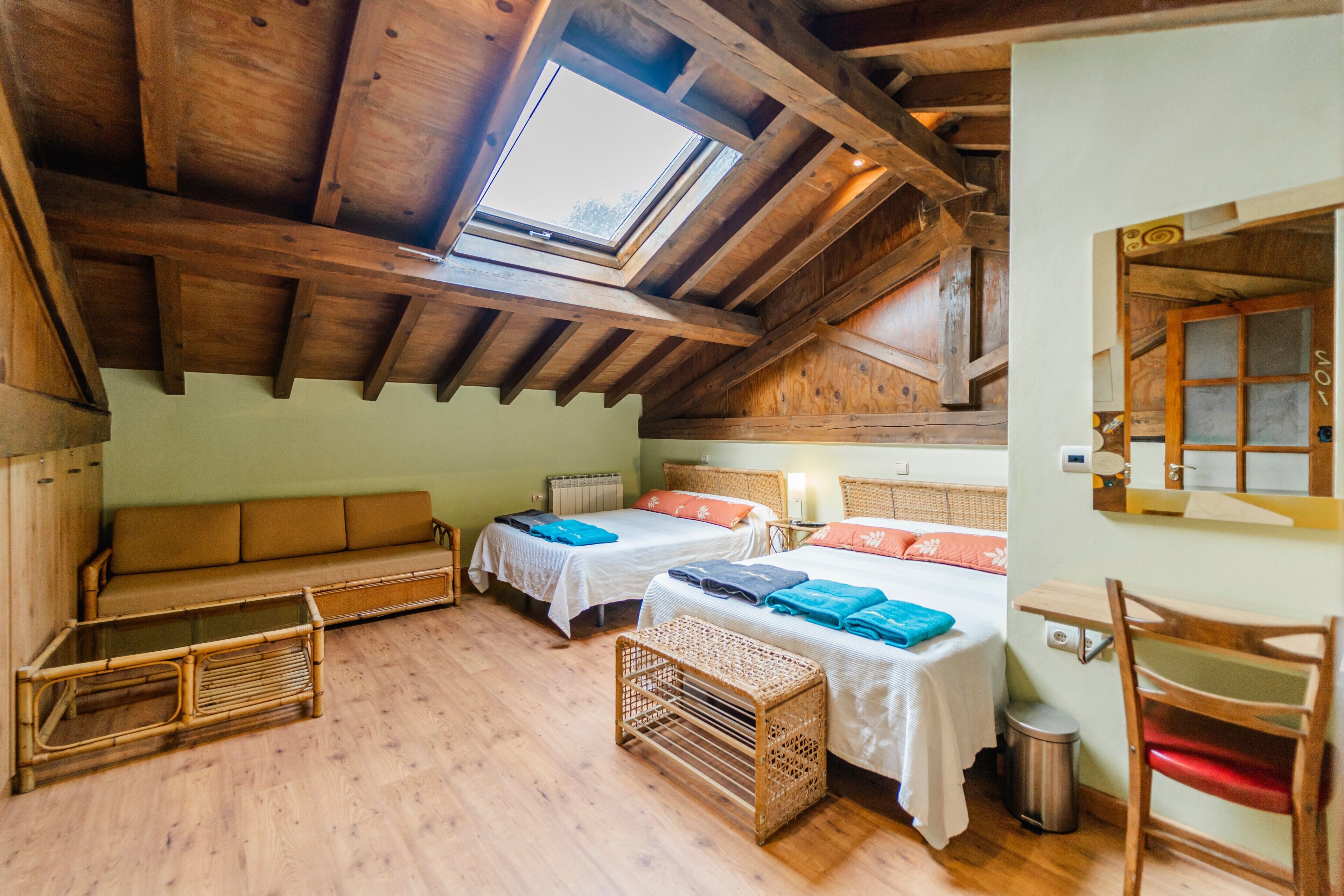 Una habitación con techo inclinado, vigas de madera, tragaluces, dos camas con ropa de cama blanca y naranja, un sofá, una cesta de mimbre y una silla.