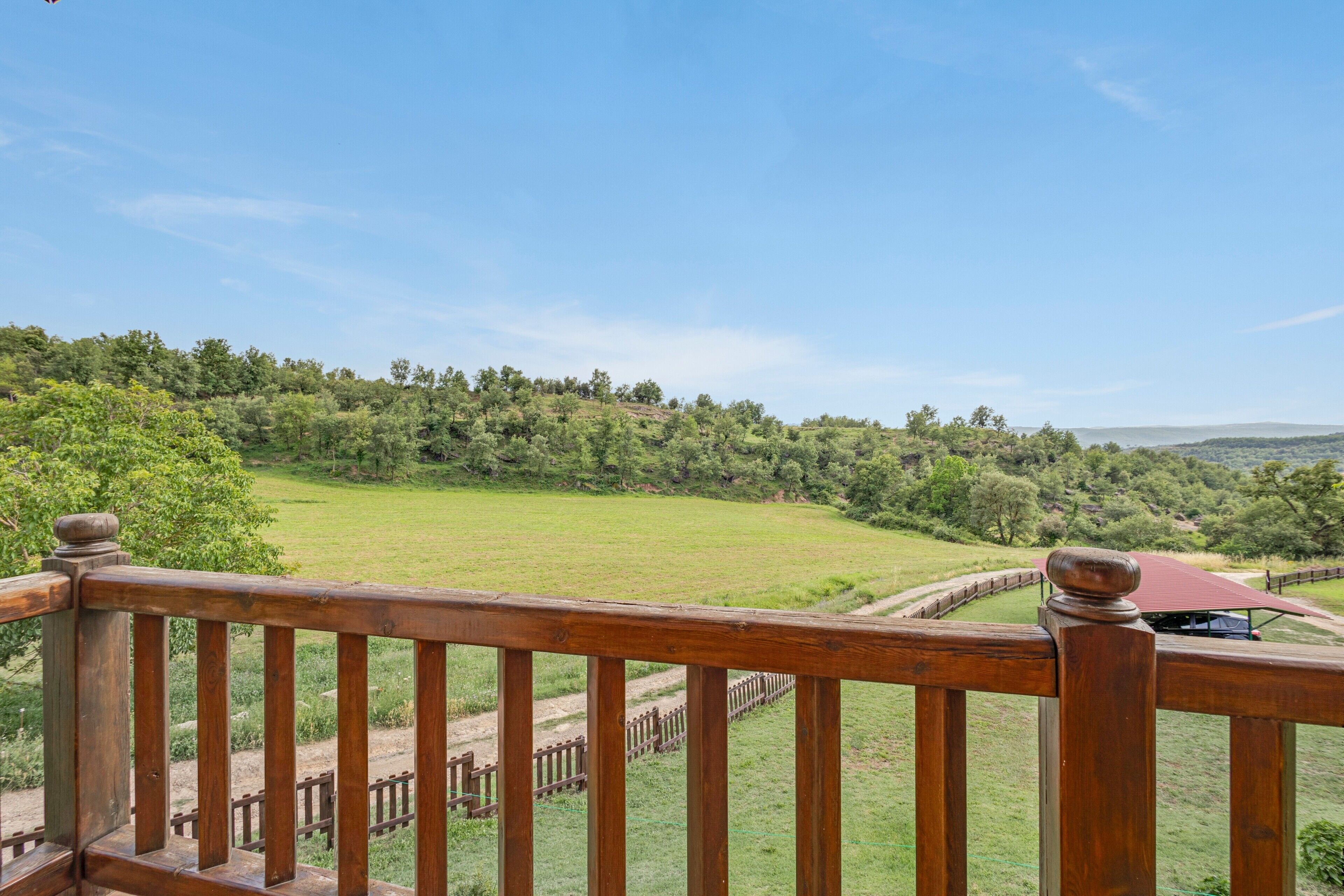 Barandilla de madera con vistas a un campo cubierto de hierba, árboles y colinas a lo lejos.