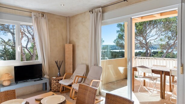 Un salon moderne doté d’un téléviseur à écran plat, de fauteuils en osier, d’une table à manger et d’un balcon offrant une vue sur les arbres et un plan d’eau.
