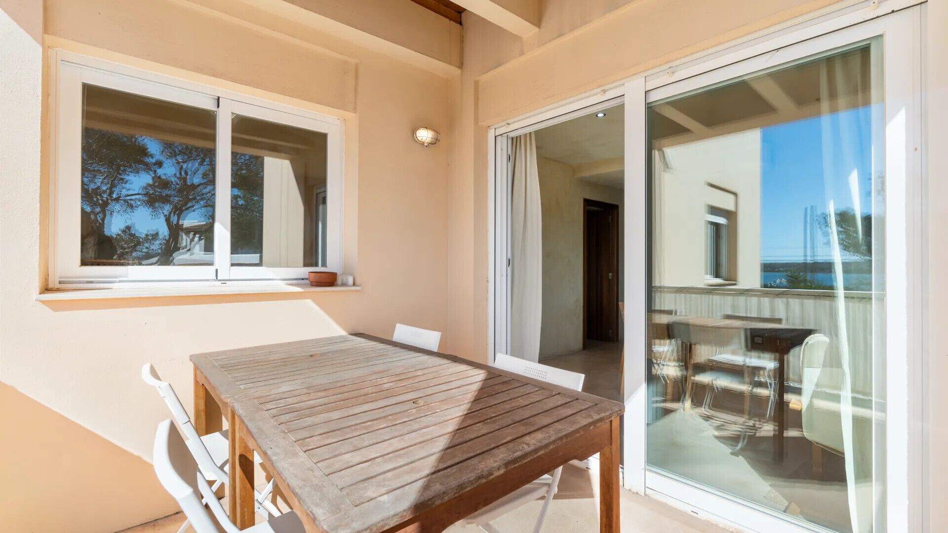 Un balcón con una mesa y sillas de madera, una ventana con vista a árboles, y un ventanal corredero que da a otra habitación.