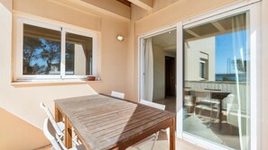 A balcony with a wooden table and chairs, a window with a view of trees, and a sliding glass door leading to another room. - Vista Azul Duplex 2 Formentera (La Savina)