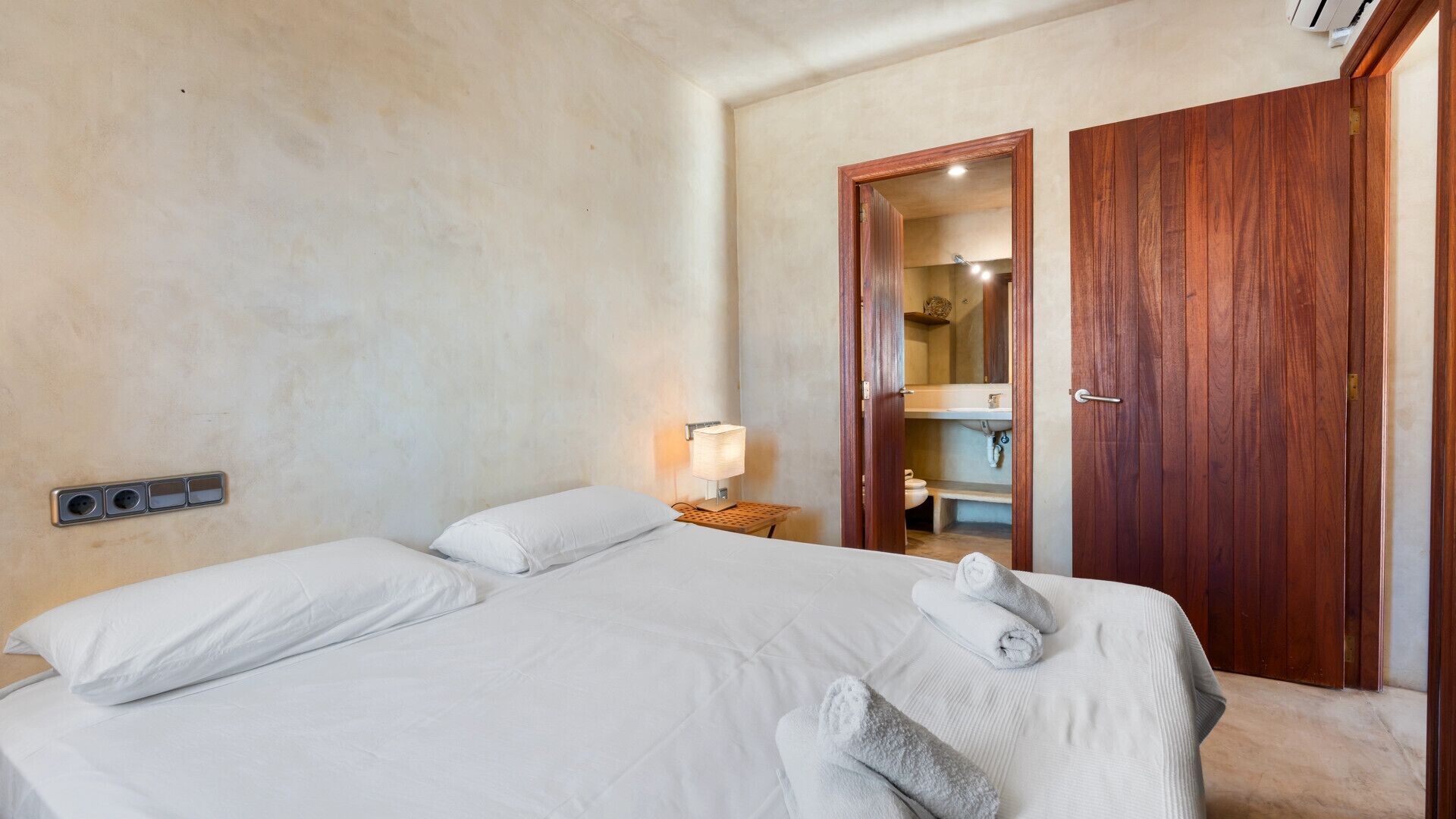 Habitación de hotel con una cama grande, una lámpara de noche, un armario de madera y un baño visible a través de una puerta abierta.