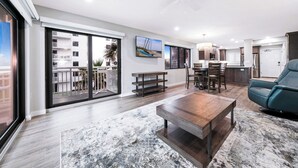 A modern living room with a large window, a ceiling fan, a dining area with a table and chairs, and a kitchen in the background. - Updated direct oceanfront corner 2/2 with a large balcony and heated beachfront pool (New Smyrna Beach)