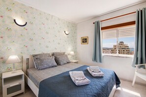 A bedroom with a bed, two bedside tables, a window with curtains, and a view of buildings.
