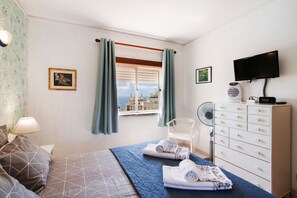 Quarto com cama, cômoda, televisão, ventilador e janela com cortinas.