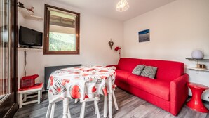 A room with a red sofa, a table with a patterned tablecloth, a television on a stand, and a window with a view of greenery. - Apartment 'Fleury' with Mountain View and Balcony (Mieussy)