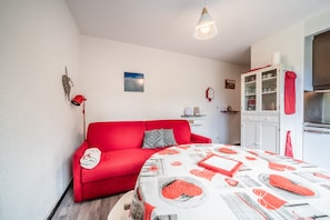 A compact living space with a red sofa, a dining table with a patterned tablecloth, a kitchen area with cabinets, and a hanging light fixture. - Apartment 'Fleury' with Mountain View and Balcony (Mieussy)