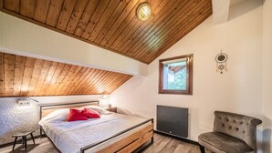 A bedroom with a wooden ceiling, a bed with a headboard, a bedside table, a chair, a TV, and a window. - Apartment 'Fleury' with Mountain View and Balcony (Mieussy)