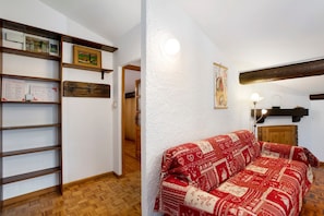 Una stanza con pavimento in legno, un divano rosso a motivi decorativi, una mensola in legno con attestati e una parete bianca dalla superficie strutturata.
