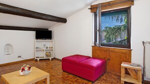 Ein Zimmer mit Holzboden, einem roten Sofa, einem kleinen Holztisch mit einer Vase, einem Fernseher auf einem weißen Regal und einem Fenster mit Blick auf Grünflächen.