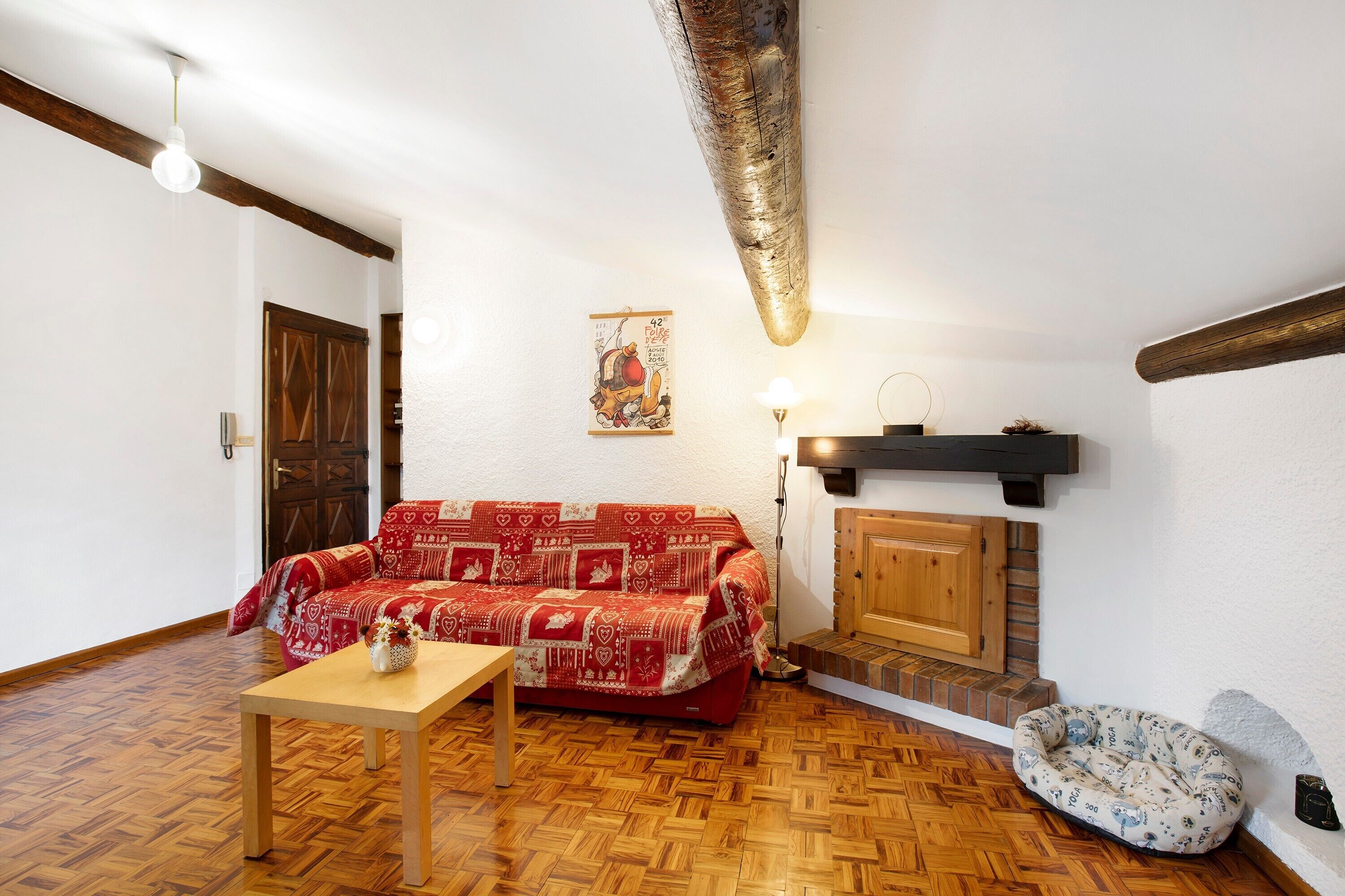 Un salon chaleureux avec un parquet en bois disposé en chevrons, un canapé rouge à motifs, une petite table en bois, un panier pour chien et une cheminée avec un manteau en bois.
