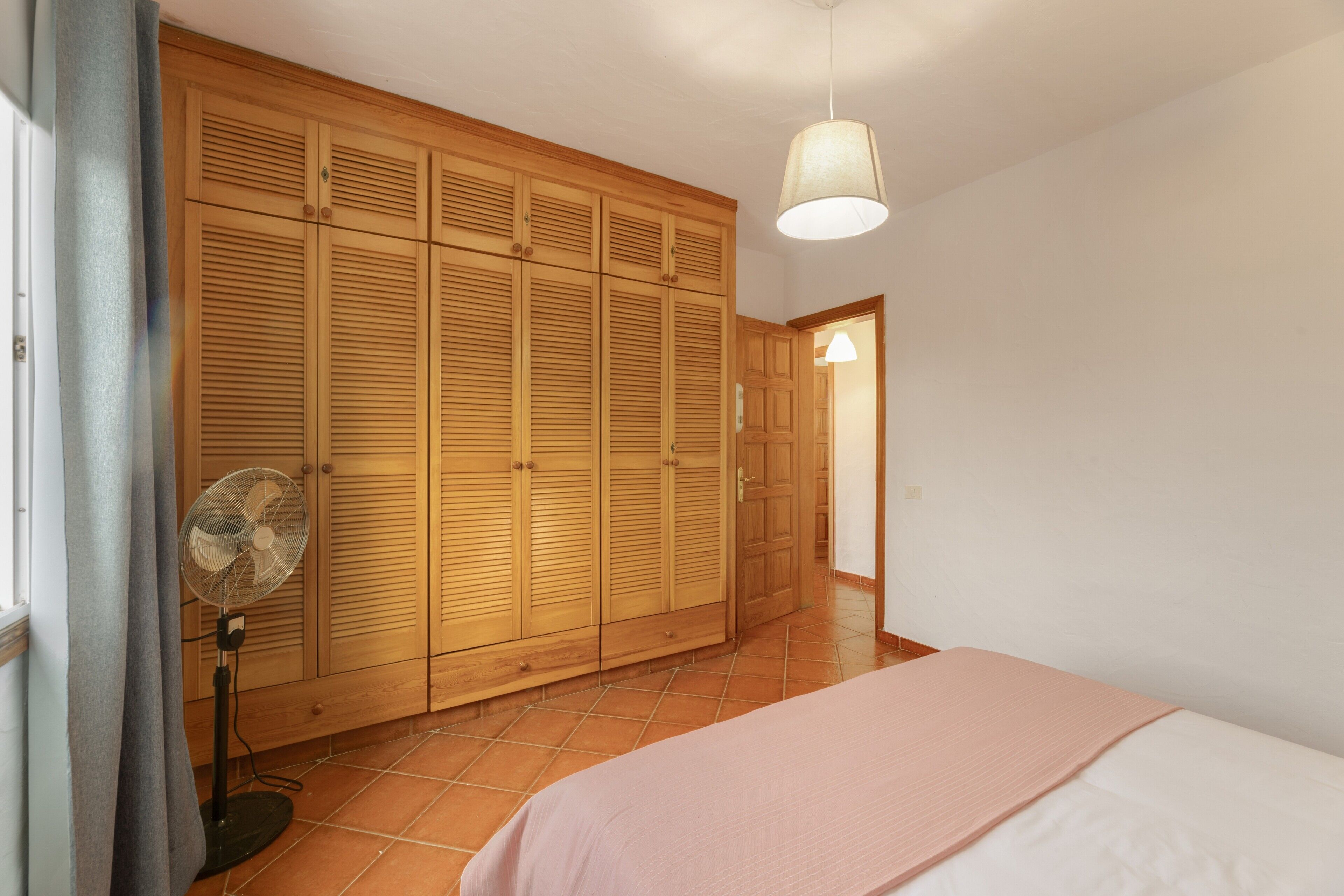 A bedroom with a bed, a wooden wardrobe, a fan, and a door leading to another room.