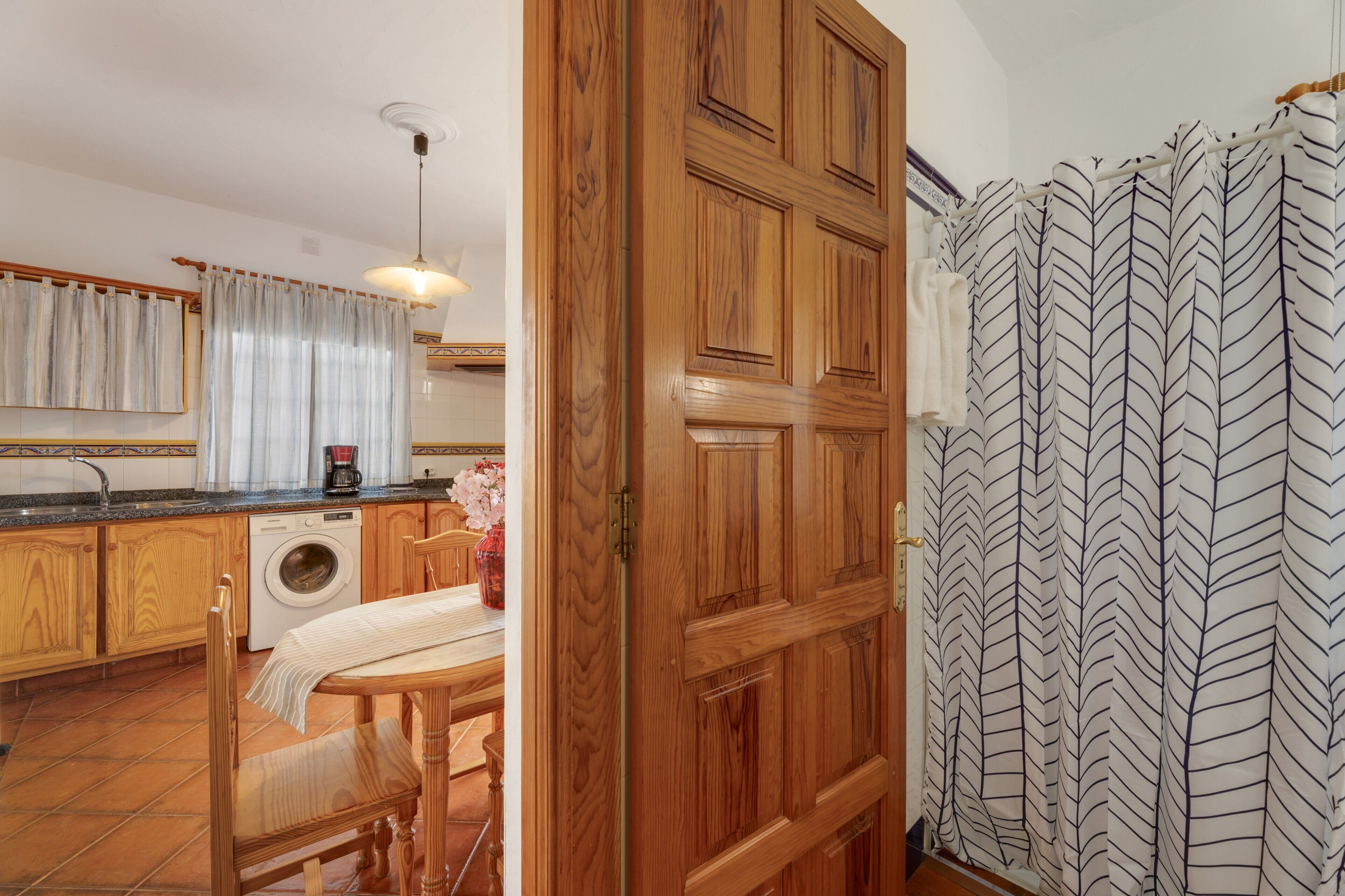 Una cocina con armarios de madera, lavadora, mesa de comedor con sillas y zona de ducha con cortinas estampadas.
