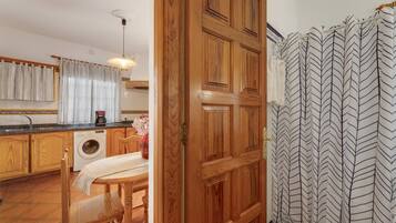 Una cocina con armarios de madera, lavadora, mesa de comedor con sillas y zona de ducha con cortinas estampadas.