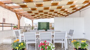 Une terrasse extérieure agrémentée d’une pergola en bois, de chaises blanches, d’une table et de plantes en pot.