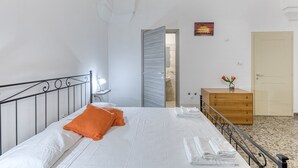 Un lit bien fait, avec du linge de lit blanc et un coussin orange, une commode en bois ornée d’un vase de fleurs, et une porte donnant sur une salle de bain.