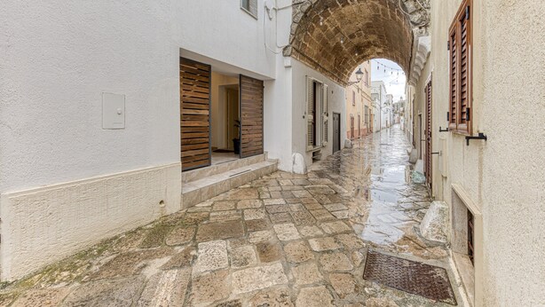 Une ruelle pavée étroite, bordée de bâtiments blancs, avec une arche et des volets en bois.