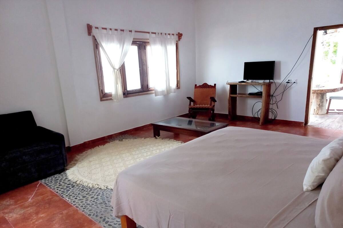 Une chambre à coucher avec un lit, une chaise, une petite table, un téléviseur et une fenêtre avec des rideaux.