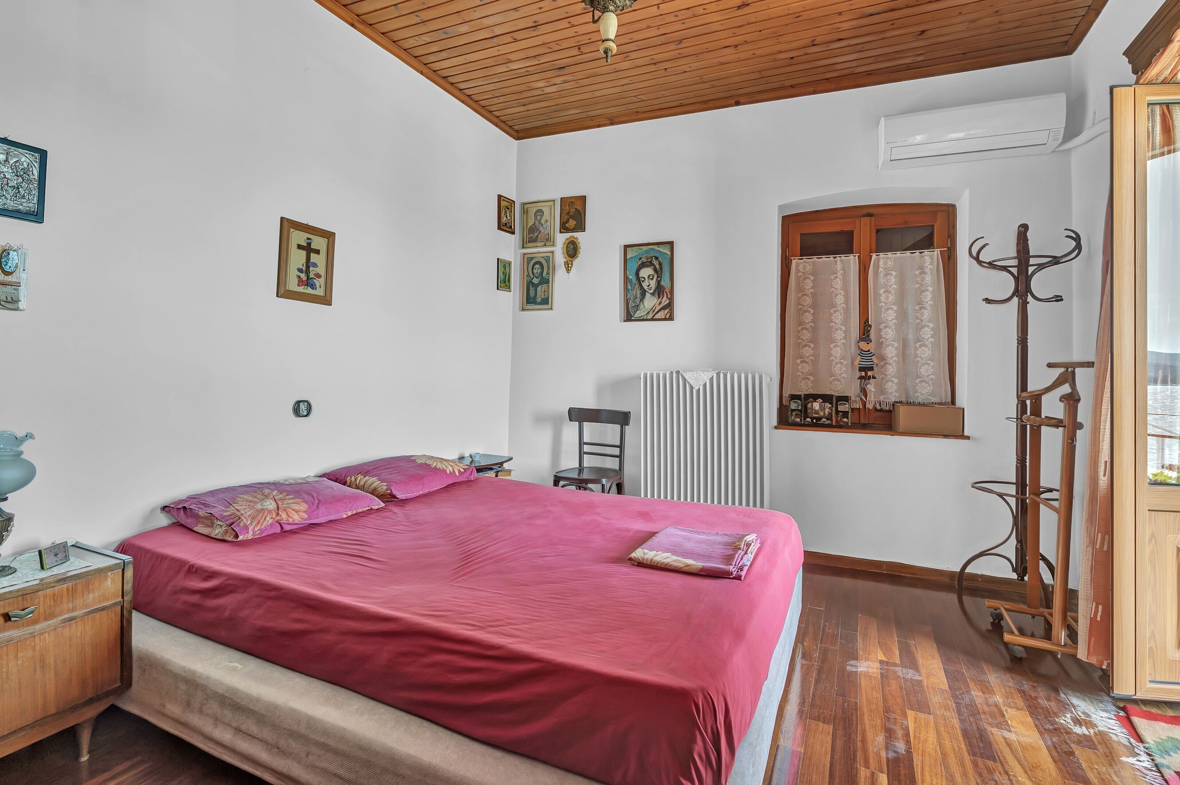 Une chambre avec un plafond en bois, un lit recouvert d’une couverture rouge, un radiateur, une fenêtre avec des rideaux et un porte-manteau.