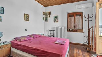 Une chambre avec un plafond en bois, un lit recouvert d’une couverture rouge, un radiateur, une fenêtre avec des rideaux et un porte-manteau.