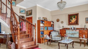 A spacious living room with a wooden staircase, a large bookshelf, a chandelier, and a dining area.